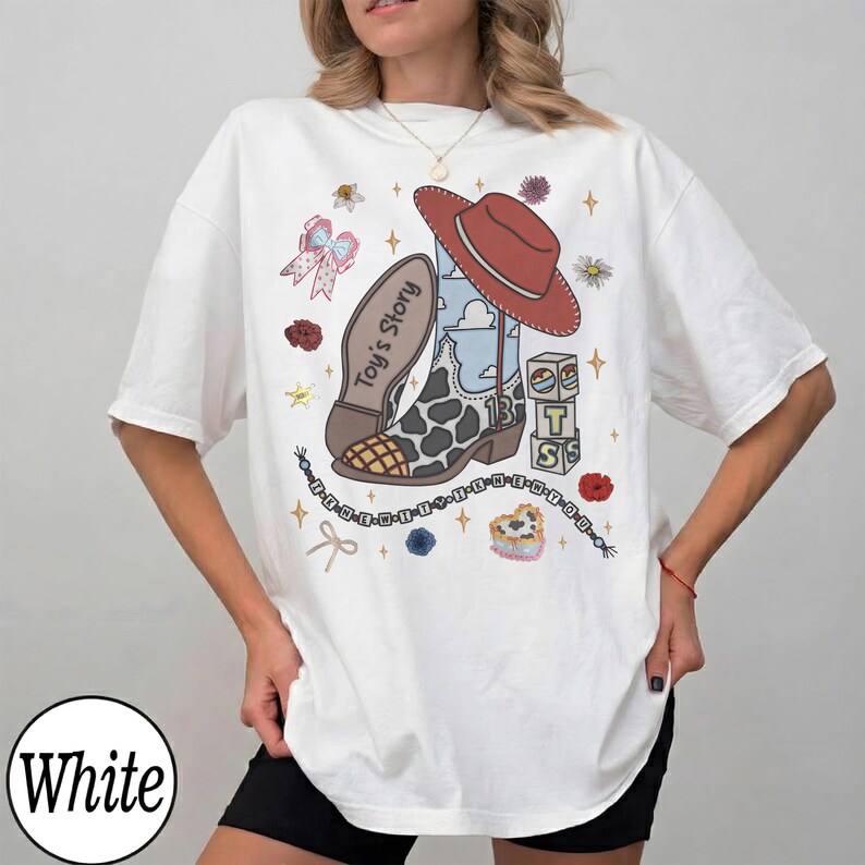 Puede incluir: Una camiseta blanca de manga corta con un estampado gr&aacute;fico de una bota de vaquero con manchas de vaca, un sombrero de vaquero rojo y bloques con las palabras "Toy's Story" y "TS". El dise&ntilde;o incluye un collar de cuentas, un lazo, flores y estrellas.