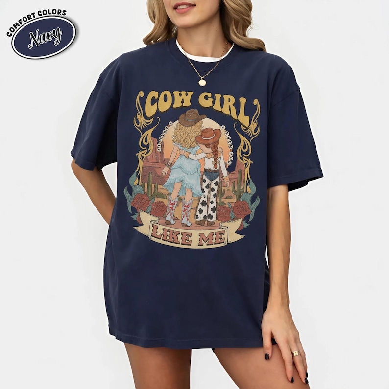 Puede incluir: Una camiseta de manga corta azul marino con un estampado gr&aacute;fico de dos figuras con sombreros de vaquero en un paisaje des&eacute;rtico con cactus y rosas. El texto "COW GIRL" aparece sobre las figuras y "LIKE ME" en una cinta inferior.