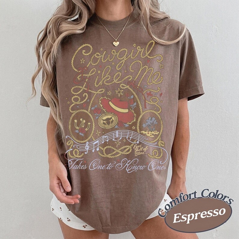 May include: A brown short-sleeve t-shirt featuring a graphic print with the text Cowgirl Like Me and Takes One to Know One. The design includes a red hat, a lasso, musical notes, and stars in yellow, red, and white ink on the front of the shirt.