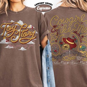 May include: An espresso-colored t-shirt featuring a front graphic with the text 'it's a Toy Story' in rope-style lettering over clouds. The back graphic displays 'Cowgirl like me' in rope text, a cowboy hat, a horseshoe, and musical notes.
