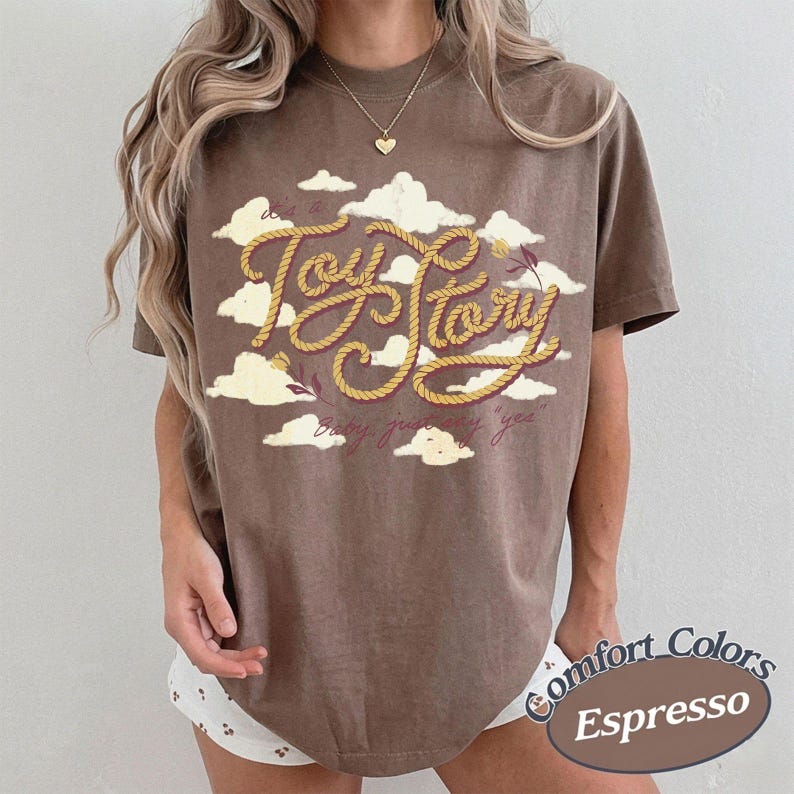 May include: A brown short-sleeve t-shirt featuring a graphic of yellow rope forming the text Toy Story, surrounded by white clouds. The shirt includes the text It's a Toy Story, baby, just say yee, and a gold heart pendant necklace is worn.