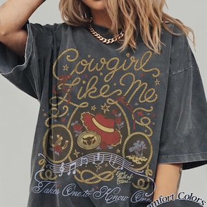 May include: A charcoal gray short-sleeve t-shirt featuring a graphic print with the text Cowgirl Like Me, Takes One to Know One, and Eyes full of Stars. The design includes a red hat, a lasso, a horseshoe, musical notes, and roses.