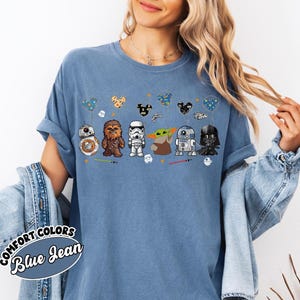May include: Blue jean colored t-shirt featuring cartoon Star Wars characters and Mickey Mouse-shaped balloons. Characters include BB-8, Chewbacca, a Stormtrooper, Grogu, R2-D2, and Darth Vader. The shirt has the words "Comfort Colors Blue Jean" on the bottom left.