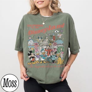 May include: Olive green t-shirt with a Disneyland graphic featuring Mickey Mouse, Minnie Mouse, Donald Duck, Goofy, and Pluto. The graphic includes the text "California Est. 1955 Disneyland" and "Where dreams come true."