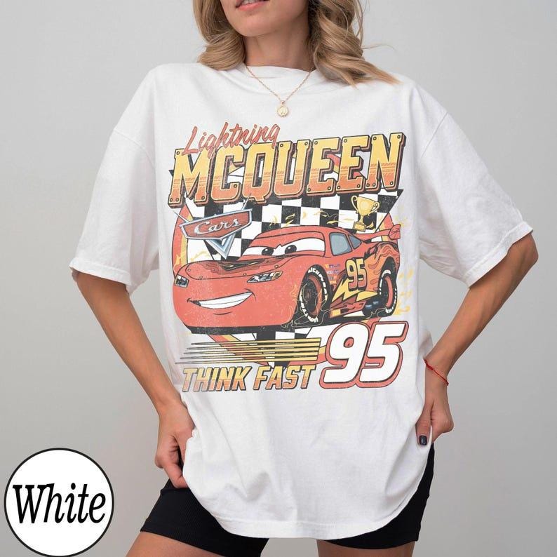 May include: White t-shirt featuring a graphic of Lightning McQueen from the Cars movie. The design includes the text "Lightning McQueen," "Cars," "Think Fast," and the number "95" in a vintage style.