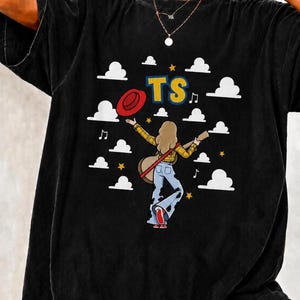 May include: A black t-shirt featuring a graphic of a person with long hair wearing a yellow plaid shirt and blue cow-print bell-bottom trousers. The person holds a guitar and a red hat. The design includes white clouds, yellow stars, and the text TS.
