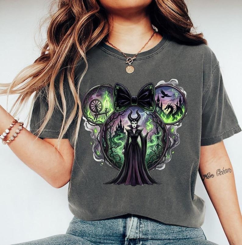 May include: Dark gray t-shirt featuring a graphic of a Disney villain, Maleficent, in a Mickey Mouse ear design. The design includes a black bow, a castle, a dragon, and green flames. The shirt is made of a soft material.
