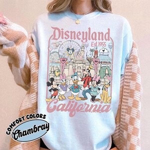 May include: Light blue Disneyland California sweatshirt with a graphic featuring Mickey Mouse, Minnie Mouse, Donald Duck, Daisy Duck, Goofy, and Pluto. The design includes the Disneyland castle, a Ferris wheel, and the text "Disneyland Est. 1955".