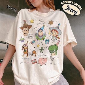 May include: An ivory short-sleeve t-shirt featuring a colourful graphic print of various animated toy characters wearing party hats. The design includes the names Woody, Buzz Lightyear, Jessie, Slinky, Hamm, and Rex, along with small illustrations of a rocket, a gift box, and stars.