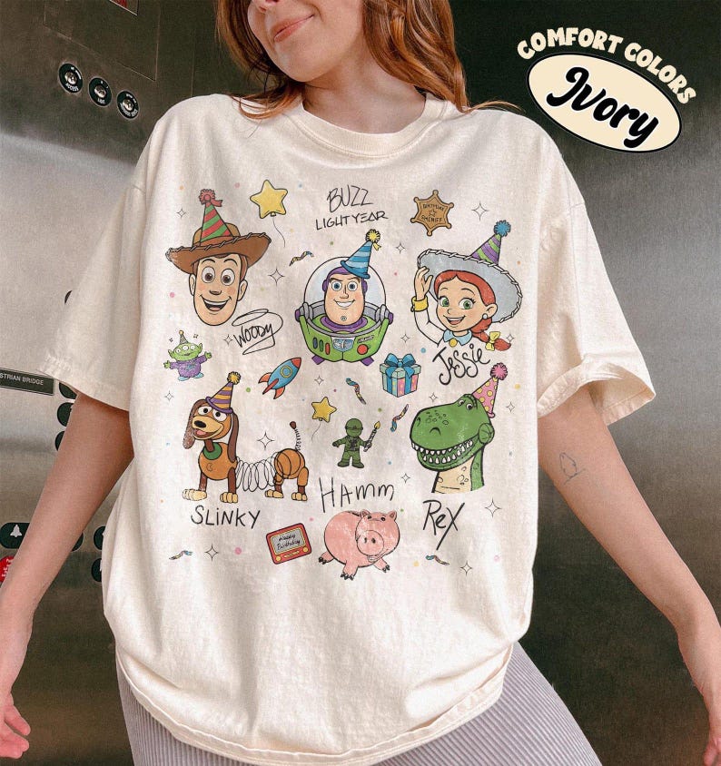 May include: An ivory short-sleeve t-shirt featuring a colourful graphic print of various animated toy characters wearing party hats. The design includes the names Woody, Buzz Lightyear, Jessie, Slinky, Hamm, and Rex, along with small illustrations of a rocket, a gift box, and stars.