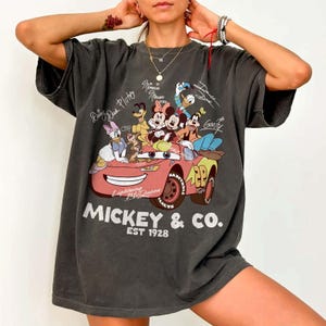 Może przedstawiać: Ciemnoszara koszulka oversize z grafiką postaci Disneya w r&oacute;żowym samochodzie. Projekt zawiera Myszkę Miki, Minnie, Kaczora Donalda i Goofy'ego, z napisem "Mickey & Co. EST 1928."
