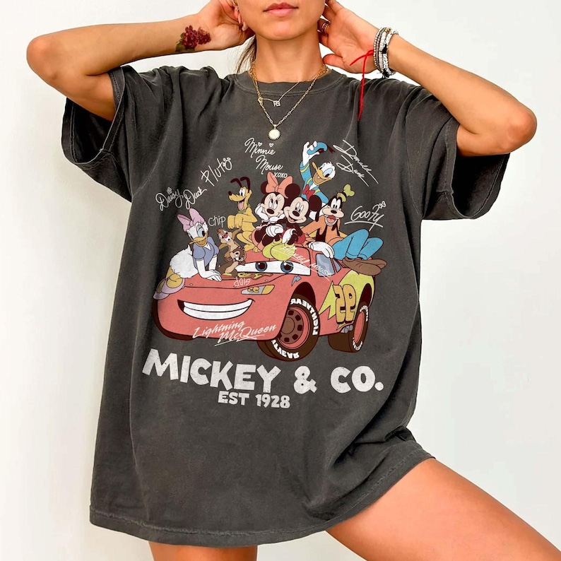 Może przedstawiać: Ciemnoszara koszulka oversize z grafiką postaci Disneya w r&oacute;żowym samochodzie. Projekt zawiera Myszkę Miki, Minnie, Kaczora Donalda i Goofy'ego, z napisem "Mickey & Co. EST 1928."