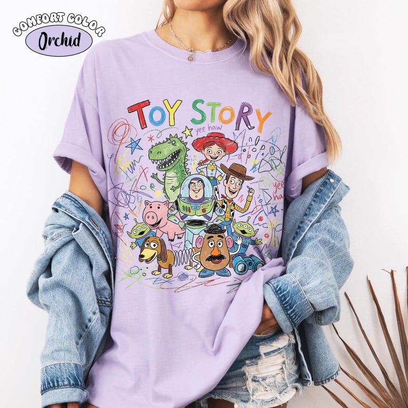 May include: A light purple t-shirt featuring a colorful graphic print of Toy Story characters including Woody, Buzz Lightyear, Jessie, Rex, Slinky Dog, Mr. Potato Head, and the Aliens, surrounded by multi-colored scribbles and the text Toy Story.