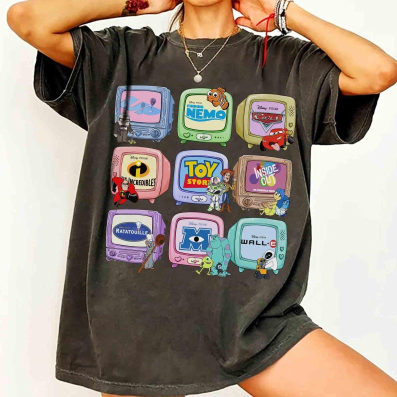 May include: Dark gray t-shirt featuring nine colorful television screens, each displaying a different Disney Pixar movie scene, including "Nemo," "Cars," and "Toy Story." The shirt has a vintage, worn look.