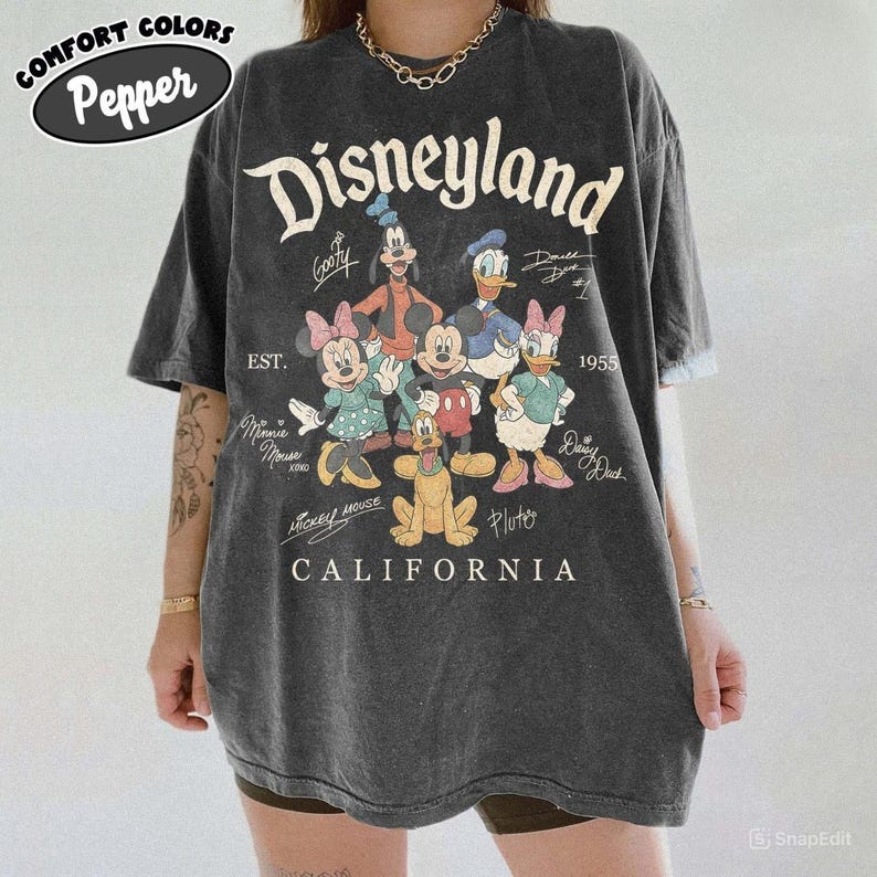 K&ouml;nnte beinhalten: Dunkelgraues T-Shirt mit dem Wort "Disneyland" in einer Vintage-Schriftart sowie Zeichentrickfiguren wie Mickey Mouse, Minnie Mouse und Donald Duck. Das Shirt enth&auml;lt auch den Text "California" und "Est. 1955".