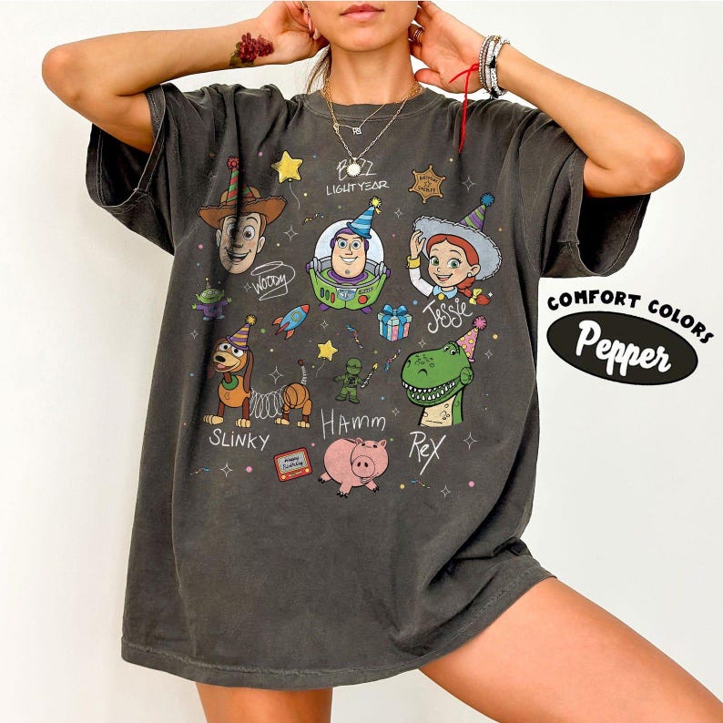 May include: A charcoal grey short-sleeve t-shirt featuring a graphic print of various animated characters including Woody, Buzz Lightyear, Jessie, Slinky, Hamm, and Rex. The design includes colourful party hats, stars, and text labels for each character.