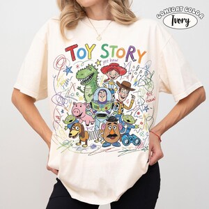May include: An ivory t-shirt featuring a colorful graphic of Toy Story characters including Woody, Buzz Lightyear, Jessie, Rex, Hamm, Slinky Dog, Mr. Potato Head, and Aliens. The design includes the text "TOY STORY" and "yee haw" with scribbled crayon-style accents.
