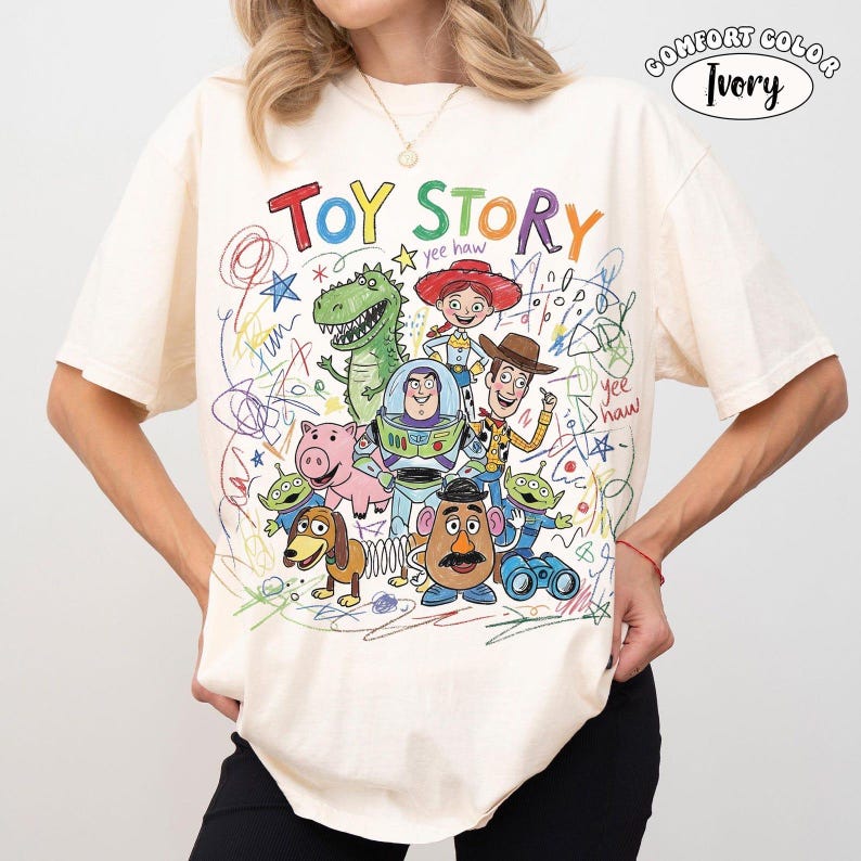 May include: An ivory t-shirt featuring a colorful graphic of Toy Story characters including Woody, Buzz Lightyear, Jessie, Rex, Hamm, Slinky Dog, Mr. Potato Head, and Aliens. The design includes the text "TOY STORY" and "yee haw" with scribbled crayon-style accents.