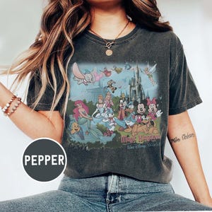 May include: Dark grey t-shirt with a colourful graphic of Disney characters, including Ariel, Dumbo, Mickey and Minnie Mouse, and various princesses. The text "Magic Kingdom, Walt Disney World" is printed below the image.