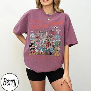 May include: Berry-coloured t-shirt featuring a Disneyland graphic. The design includes Mickey Mouse, Donald Duck, Goofy, and other Disney characters in front of a castle. The shirt has a crew neck and short sleeves.