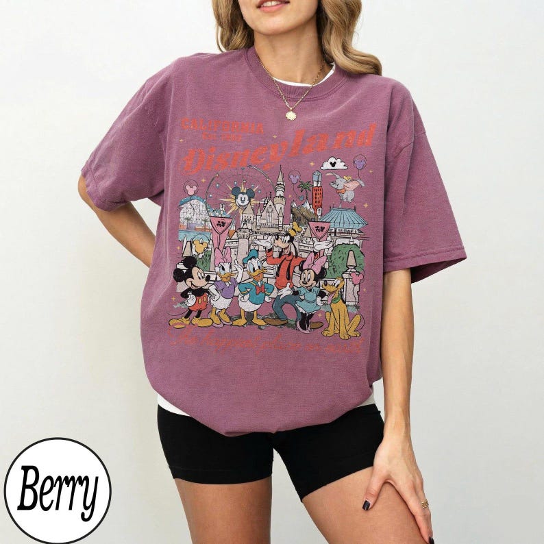 May include: Berry-coloured t-shirt featuring a Disneyland graphic. The design includes Mickey Mouse, Donald Duck, Goofy, and other Disney characters in front of a castle. The shirt has a crew neck and short sleeves.