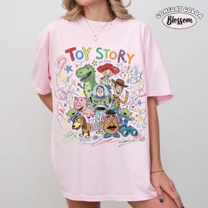 May include: A light pink t-shirt featuring a colorful graphic print of Toy Story characters including Buzz Lightyear, Woody, Jessie, Rex, Hamm, Mr. Potato Head, and aliens. The design includes scribbled crayon-style drawings and the text Toy Story.