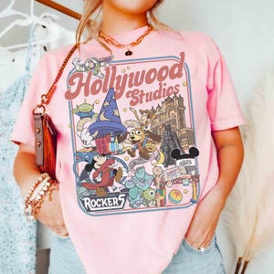 K&ouml;nnte beinhalten: Hellrosa T-Shirt mit einer farbenfrohen Grafik, die Hollywood Studios Charaktere und Attraktionen zeigt. Das Design umfasst Mickey Mouse, Buzz Lightyear und das Hollywood Studios Logo. Das Wort "Rockers" ist ebenfalls sichtbar.