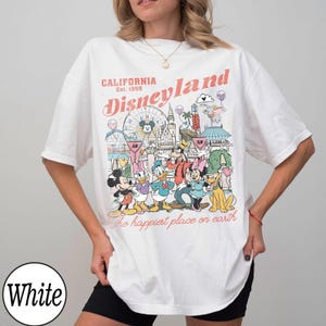 May include: White t-shirt with a Disneyland graphic. The design includes the text "California Disneyland Est. 1955" and "The happiest place on earth". The graphic features cartoon characters and park landmarks.