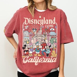 May include: Red t-shirt featuring a Disneyland graphic. The design includes the words "Disneyland Est 1955 California" and cartoon characters Mickey Mouse, Minnie Mouse, Donald Duck, Daisy Duck, Goofy, and Pluto. The shirt has a vintage, distressed look.