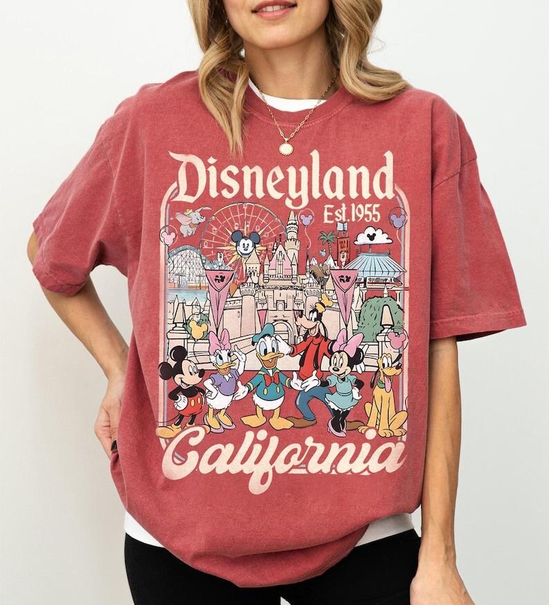 May include: Red t-shirt featuring a Disneyland graphic. The design includes the words "Disneyland Est 1955 California" and cartoon characters Mickey Mouse, Minnie Mouse, Donald Duck, Daisy Duck, Goofy, and Pluto. The shirt has a vintage, distressed look.