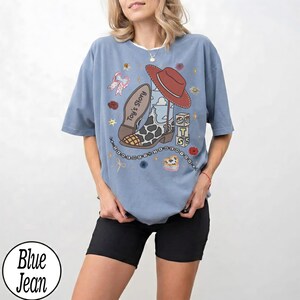 Puede incluir: Una camiseta de color azul vaquero con un estampado gr&aacute;fico de una bota de vaquero, un sombrero rojo y bloques con el texto Toy's Story. El dise&ntilde;o incluye peque&ntilde;os elementos decorativos como flores, estrellas y una cadena de cuentas alrededor.