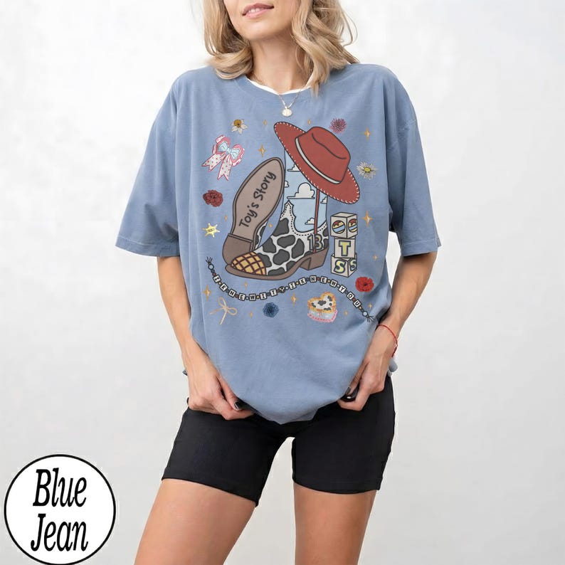 Puede incluir: Una camiseta de color azul vaquero con un estampado gr&aacute;fico de una bota de vaquero, un sombrero rojo y bloques con el texto Toy's Story. El dise&ntilde;o incluye peque&ntilde;os elementos decorativos como flores, estrellas y una cadena de cuentas alrededor.