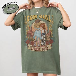 Puede incluir: Una camiseta de manga corta color verde musgo con un estampado gr&aacute;fico de dos figuras con sombreros y botas de vaquero en un paisaje des&eacute;rtico con cactus y rosas. El texto dice COW GIRL LIKE ME, con texto m&aacute;s peque&ntilde;o alrededor.