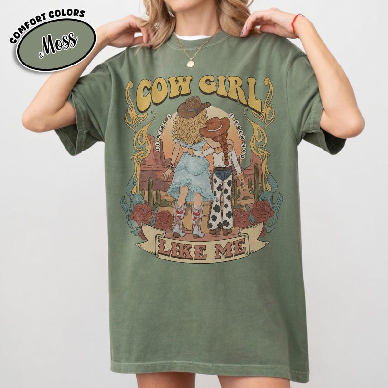 Puede incluir: Una camiseta de manga corta color verde musgo con un estampado gr&aacute;fico de dos figuras con sombreros y botas de vaquero en un paisaje des&eacute;rtico con cactus y rosas. El texto dice COW GIRL LIKE ME, con texto m&aacute;s peque&ntilde;o alrededor.