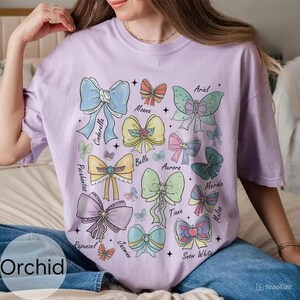 Peut inclure: Un t-shirt lilas avec une collection de n&oelig;uds color&eacute;s, chacun portant le nom d'une princesse Disney. Les n&oelig;uds sont dans diverses teintes pastel, dont le bleu, le rose, le jaune et le vert. Le mot "Orchid" est imprim&eacute; en bas &agrave; gauche.