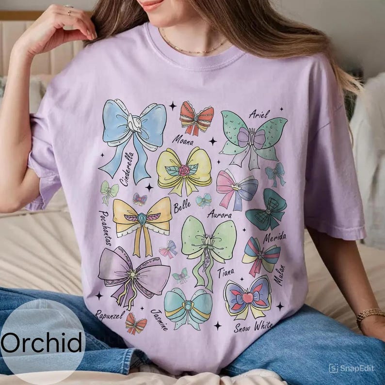 Peut inclure: Un t-shirt lilas avec une collection de n&oelig;uds color&eacute;s, chacun portant le nom d'une princesse Disney. Les n&oelig;uds sont dans diverses teintes pastel, dont le bleu, le rose, le jaune et le vert. Le mot "Orchid" est imprim&eacute; en bas &agrave; gauche.