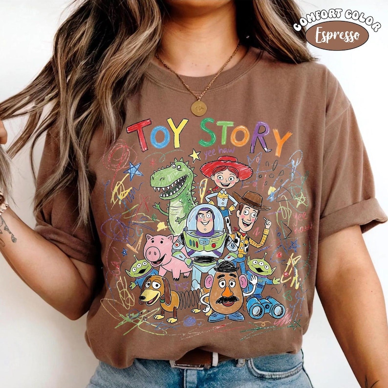 May include: An espresso brown t-shirt featuring a colorful graphic of Toy Story characters including Woody, Buzz Lightyear, Jessie, Rex, Hamm, Slinky Dog, Mr. Potato Head, and Aliens, surrounded by multi-colored scribbles and the text Toy Story.