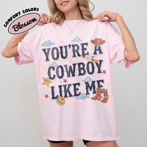 May include: A light pink short-sleeve t-shirt featuring the dark blue text "YOU'RE A COWBOY LIKE ME" with graphic illustrations of a cowboy hat, a rocket ship, a sheriff star badge, cowboy boots, a ball, clouds, and small stars.