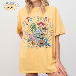 May include: A mustard yellow short-sleeve t-shirt featuring a colorful graphic print of Toy Story characters including Woody, Buzz Lightyear, Jessie, Rex, Hamm, Slinky Dog, Mr. Potato Head, and aliens, surrounded by scribbled drawings and text.