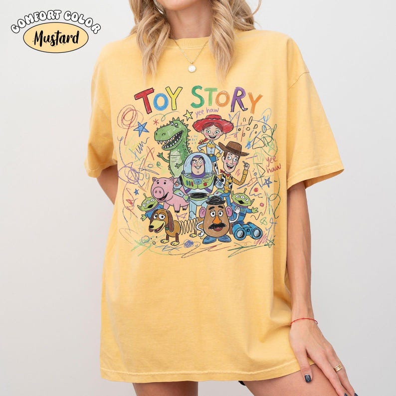 May include: A mustard yellow short-sleeve t-shirt featuring a colorful graphic print of Toy Story characters including Woody, Buzz Lightyear, Jessie, Rex, Hamm, Slinky Dog, Mr. Potato Head, and aliens, surrounded by scribbled drawings and text.