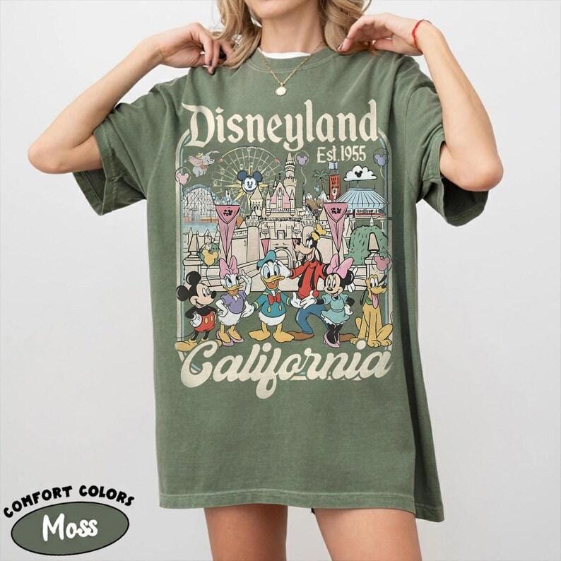 May include: Moss green t-shirt featuring a Disneyland graphic with Mickey Mouse, Donald Duck, and other characters. The graphic includes the text "Disneyland California Est. 1955". The shirt is a Comfort Colors brand.