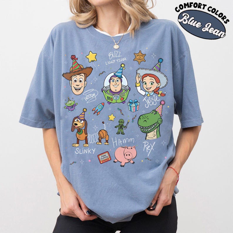 May include: A blue jean coloured t-shirt featuring a graphic print of Toy Story characters including Woody, Buzz Lightyear, Jessie, Slinky Dog, Rex, and Hamm wearing party hats. The design includes stars, confetti, and text labels for each character.