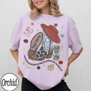 Puede incluir: Una camiseta de color morado claro con un estampado gr&aacute;fico de una bota de vaquero con patr&oacute;n de vaca, un sombrero rojo y bloques con el texto Toy's Story. El dise&ntilde;o incluye un cord&oacute;n de cuentas con el texto I KNEW IT YOU KNEW IT YOU.