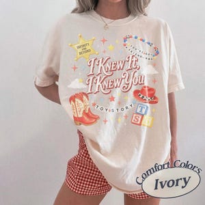 以下が含まれることがあります： 「I KNEW IT, I KNEW YOU」および「TOY STORY」の文字がプリントされたアイボリーの半袖Tシャツ。デザインには、黄色い星、ビーズのブレスレット、赤いカウボーイブーツ、赤いカウボーイハット、そしてT、S、5と書かれたアルファベットブロックが含まれています。