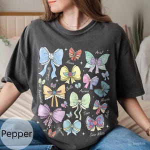 Peut inclure: T-shirt gris fonc&eacute; avec des illustrations de n&oelig;uds color&eacute;s, chacun &eacute;tiquet&eacute; avec le nom d'une princesse. Les n&oelig;uds sont de diff&eacute;rentes couleurs, dont le bleu, le jaune, le rose et le vert. Le mot "Pepper" est imprim&eacute; en bas &agrave; gauche.