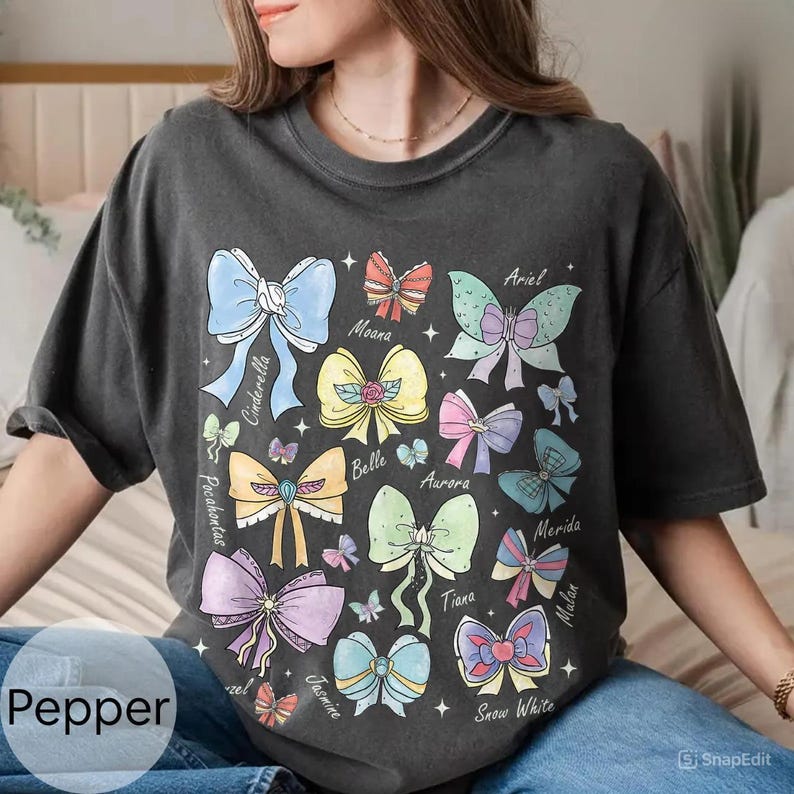 Peut inclure: T-shirt gris fonc&eacute; avec des illustrations de n&oelig;uds color&eacute;s, chacun &eacute;tiquet&eacute; avec le nom d'une princesse. Les n&oelig;uds sont de diff&eacute;rentes couleurs, dont le bleu, le jaune, le rose et le vert. Le mot "Pepper" est imprim&eacute; en bas &agrave; gauche.
