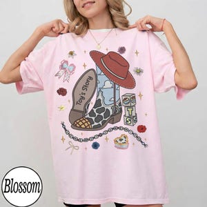 Puede incluir: Una camiseta rosa claro con un estampado gr&aacute;fico de una bota de vaquero con patr&oacute;n de manchas de vaca, un sombrero rojo y el texto Toy's Story. El dise&ntilde;o incluye peque&ntilde;as ilustraciones de un lazo, bloques de letras, flores y cuentas.