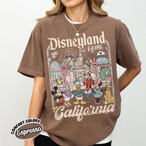May include: Brown t-shirt with a Disneyland California graphic featuring Mickey Mouse, Minnie Mouse, Donald Duck, Goofy, and Pluto. The shirt has the words "Disneyland Est. 1955" and "California" printed on it. The shirt is labeled "Comfort Colors Espresso".