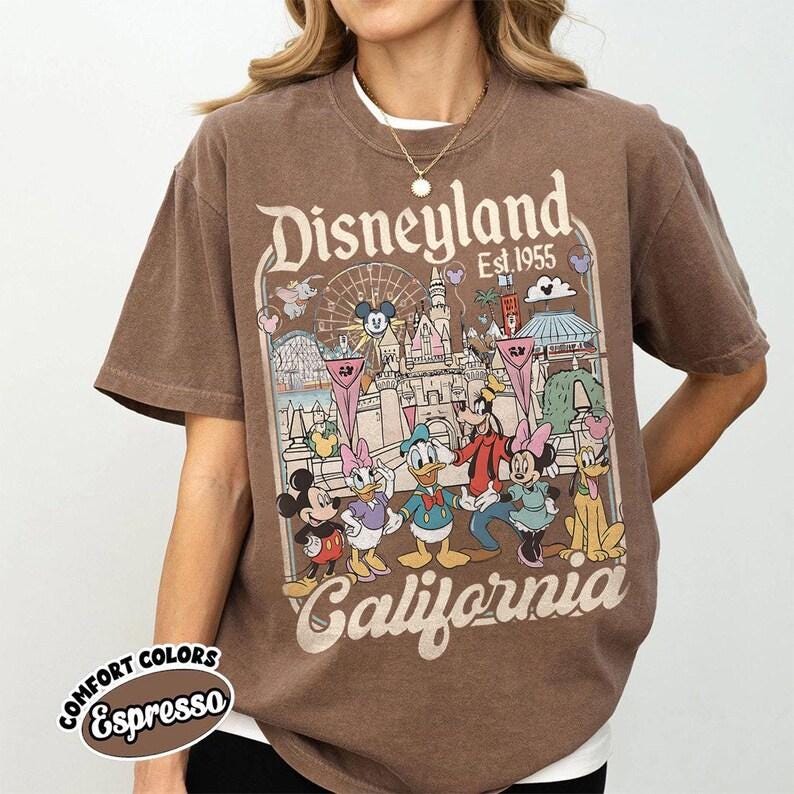 May include: Brown t-shirt with a Disneyland California graphic featuring Mickey Mouse, Minnie Mouse, Donald Duck, Goofy, and Pluto. The shirt has the words "Disneyland Est. 1955" and "California" printed on it. The shirt is labeled "Comfort Colors Espresso".
