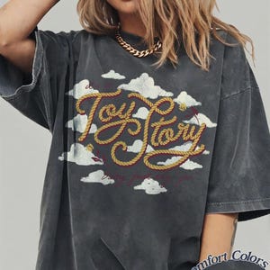 May include: A charcoal grey oversized t-shirt featuring a graphic of white clouds and the text Toy Story written in a yellow rope-style font. Below the main text, smaller red cursive text reads baby just say yes. A Comfort Colors Pepper label is visible.